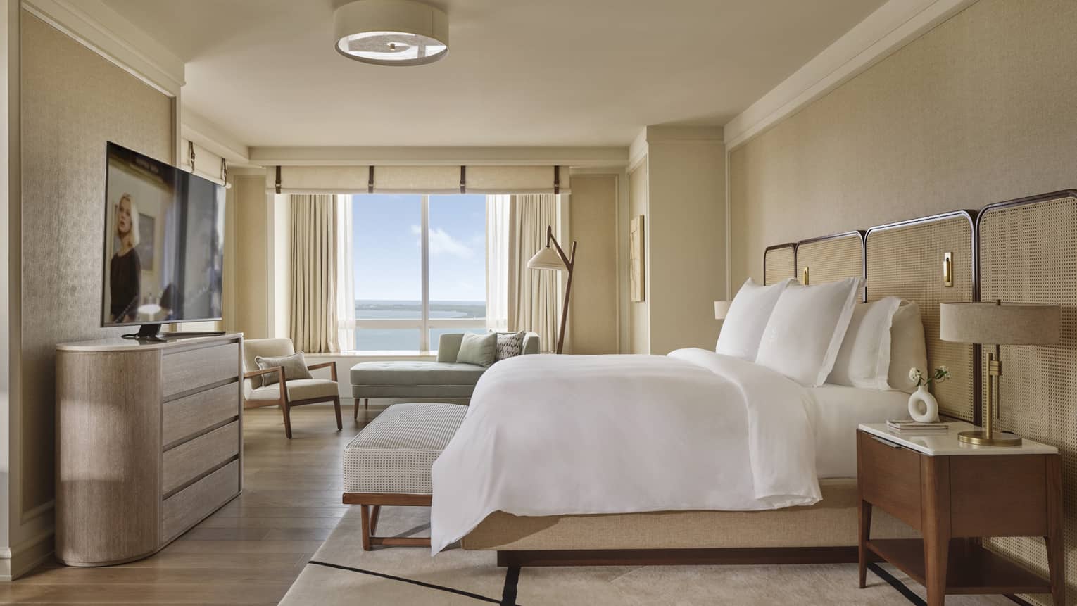 Hotel suite bedroom with king bed, bench, dresser, TV and large sea-view window