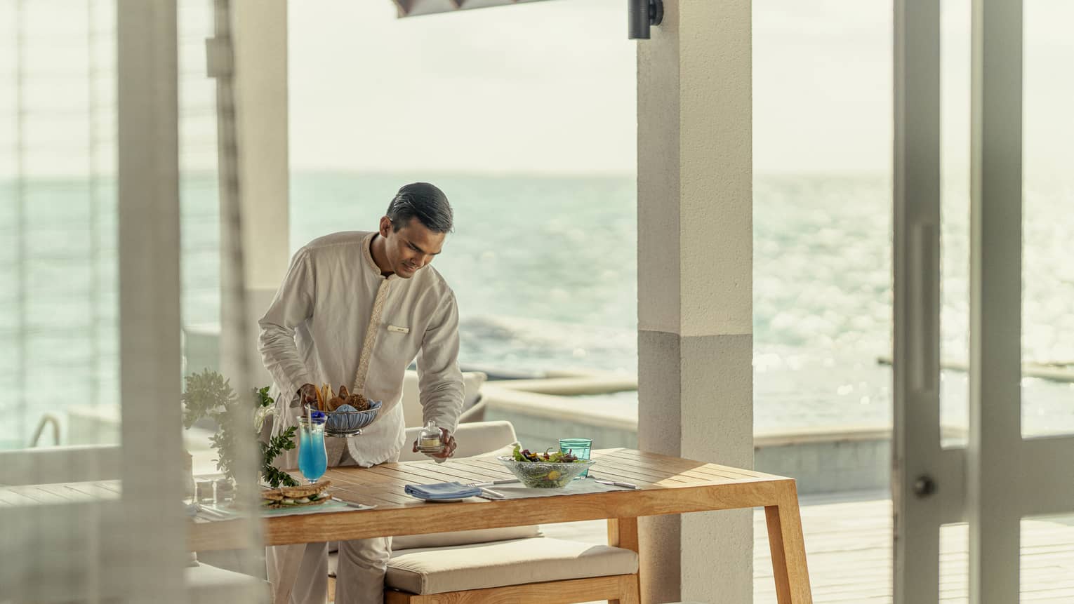 Male staff member sets table for In-Villa Dining, ocean in background