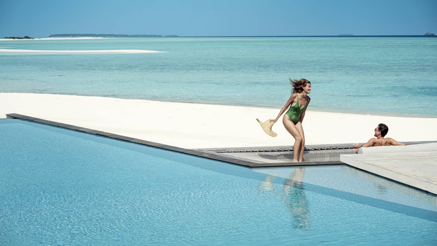 Woman and green one-piece swimsuit relaxes poolside with her male partner in the infinity pool, the vast blue waters of the lagoon behind them