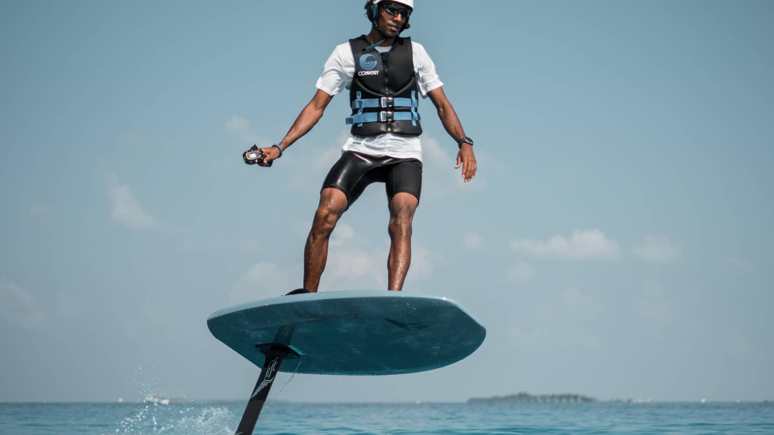 In a helmet and life jacket, an adventurer stands on an eFoil using a handheld control to propel the board out of the water.