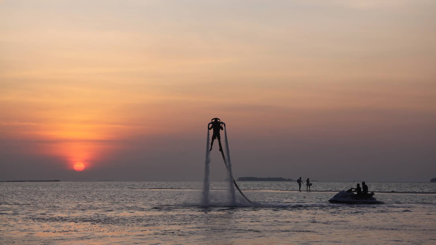 Silhouette of someone with X Jetpack suspended in air over water at sunset