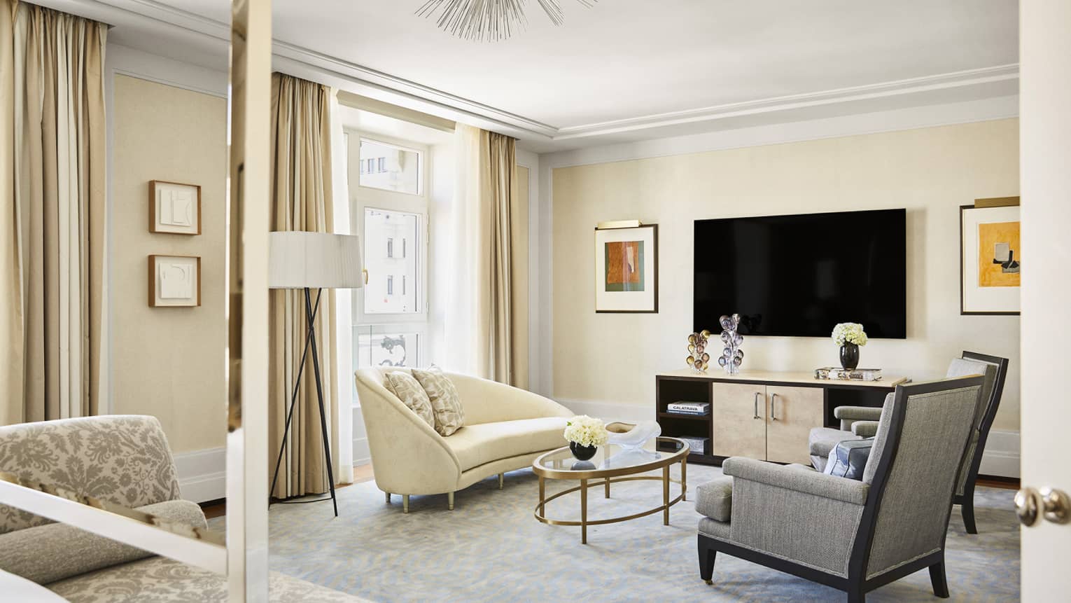 Luxury suite living area with cream chaise, grey armchairs, glass coffee table, wall-mounted TV, art pieces and floor-to-ceiling windows with beige curtains