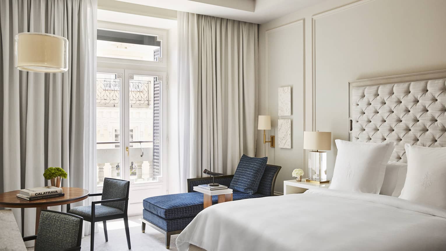 Luxury hotel bedroom with a king bed, tufted headboard, blue chaise lounge, round table with books and French doors with balcony view