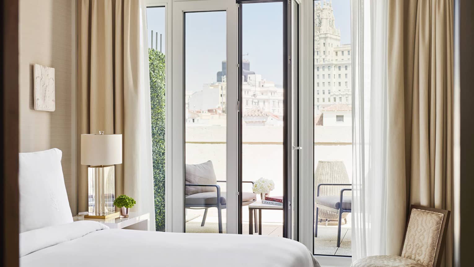 Luxury hotel bedroom with a king bed, small armchair, bedside table with lamp and a view of an outdoor terrace with seating through glass doors