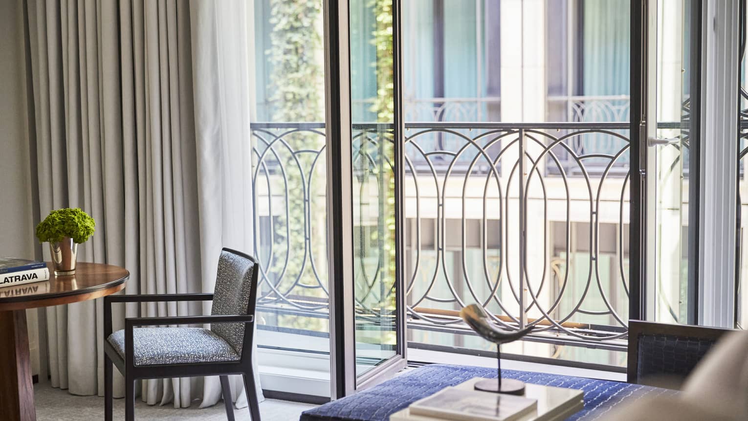 Luxury hotel room interior with a small round table, green plant, chair, horn sculpture, books and decorative railing outside a large window with curtains