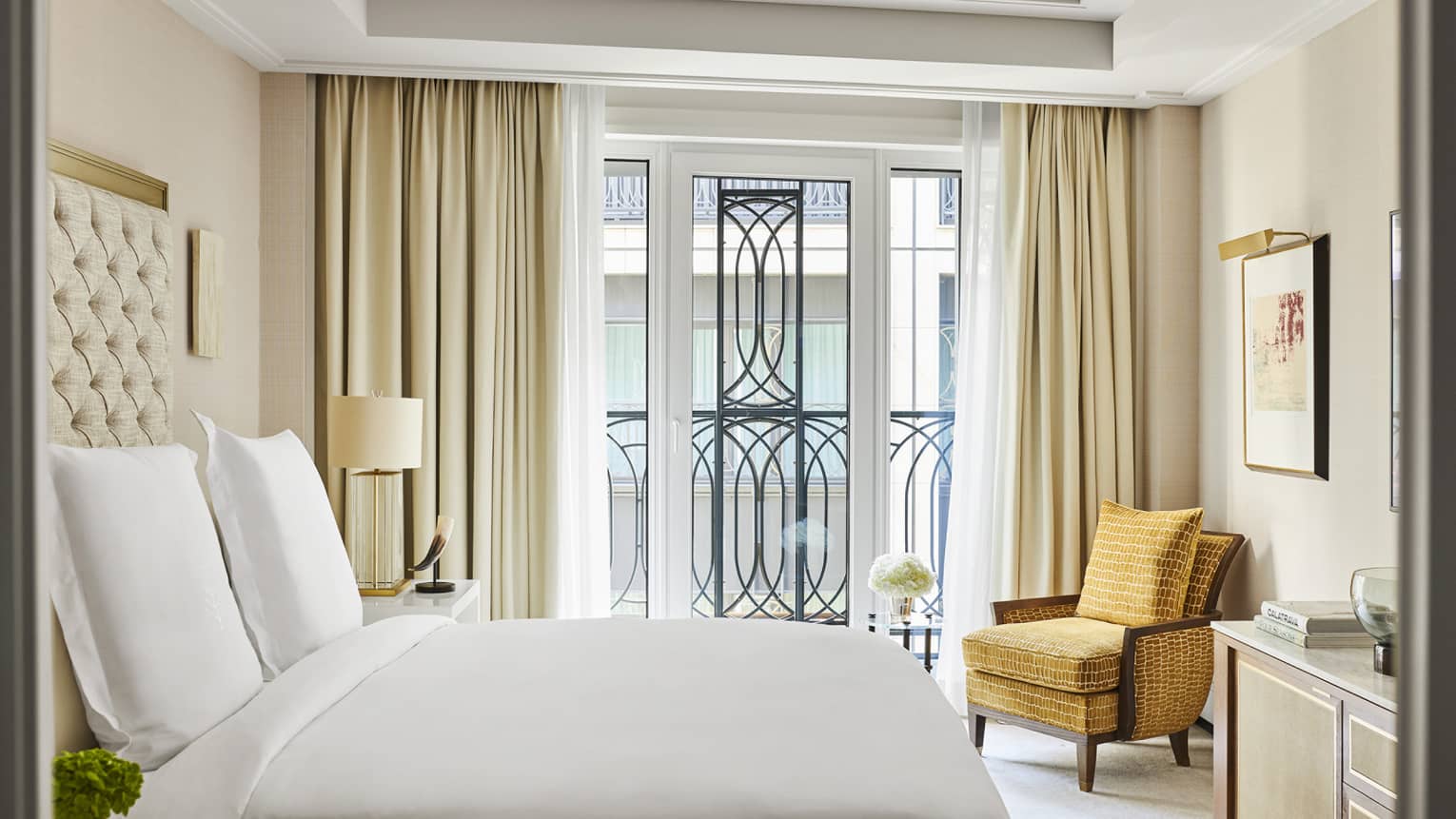 Luxury hotel bedroom with a king bed with tufted headboard, mustard armchair, bedside lamp, framed artwork and large windows featuring ornate balcony railing