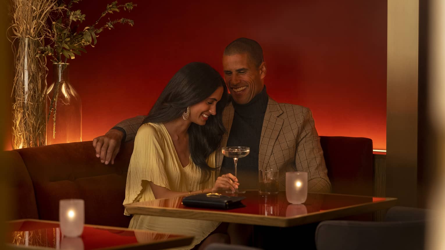 Two guests sitting together at a dining table with a candle on it, holding a glass of wine.