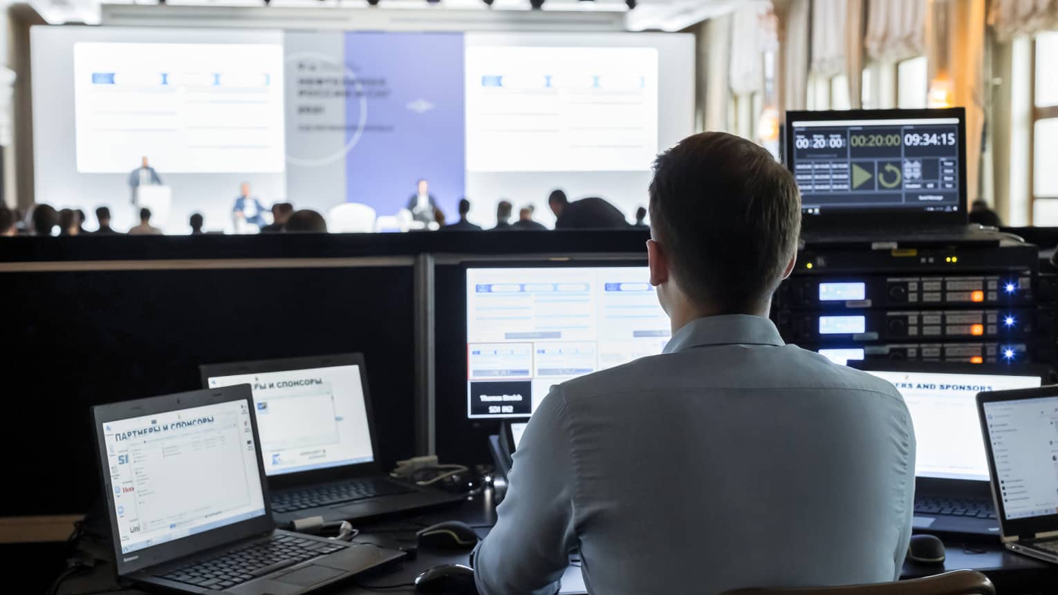 A man sits in front of several screens, projecting a slideshow in a large conference room.