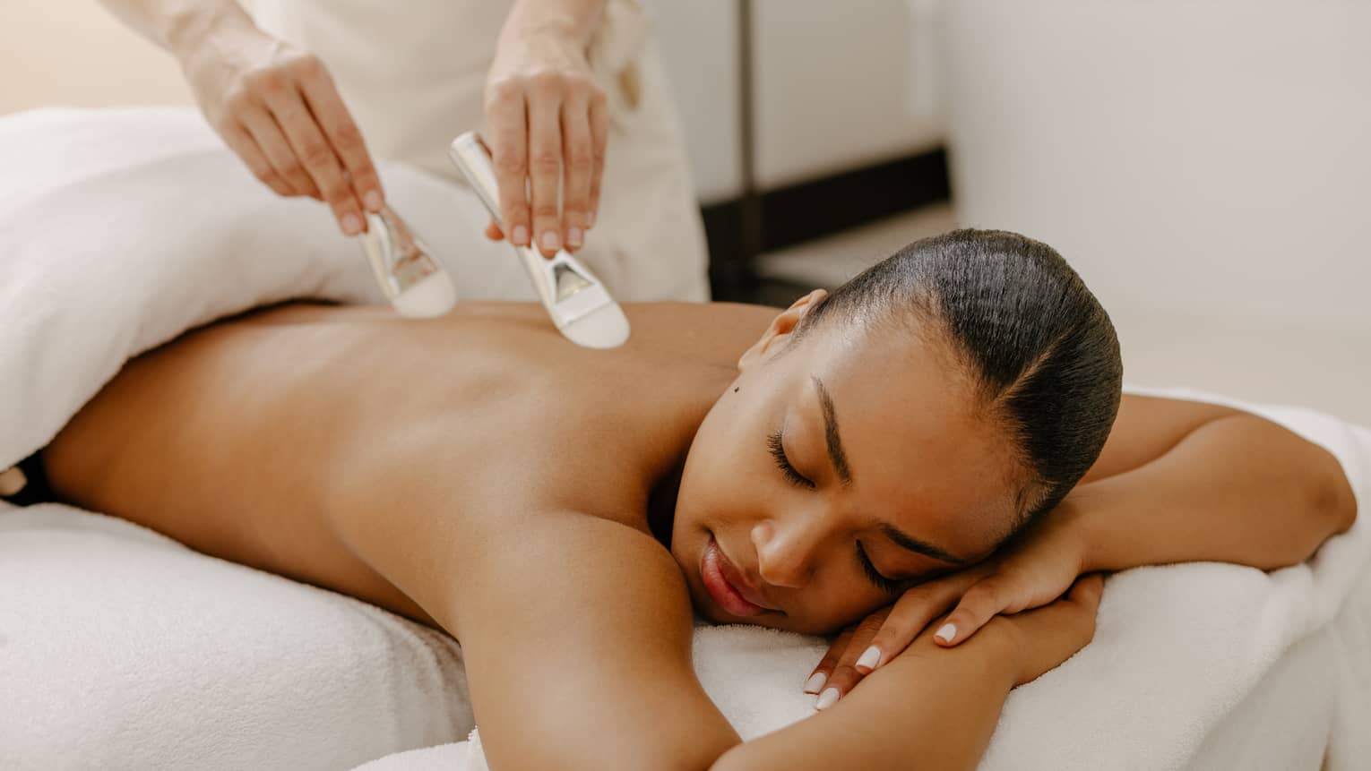 Two brushes massage a woman's back as she lay on a spa table covered with white linens