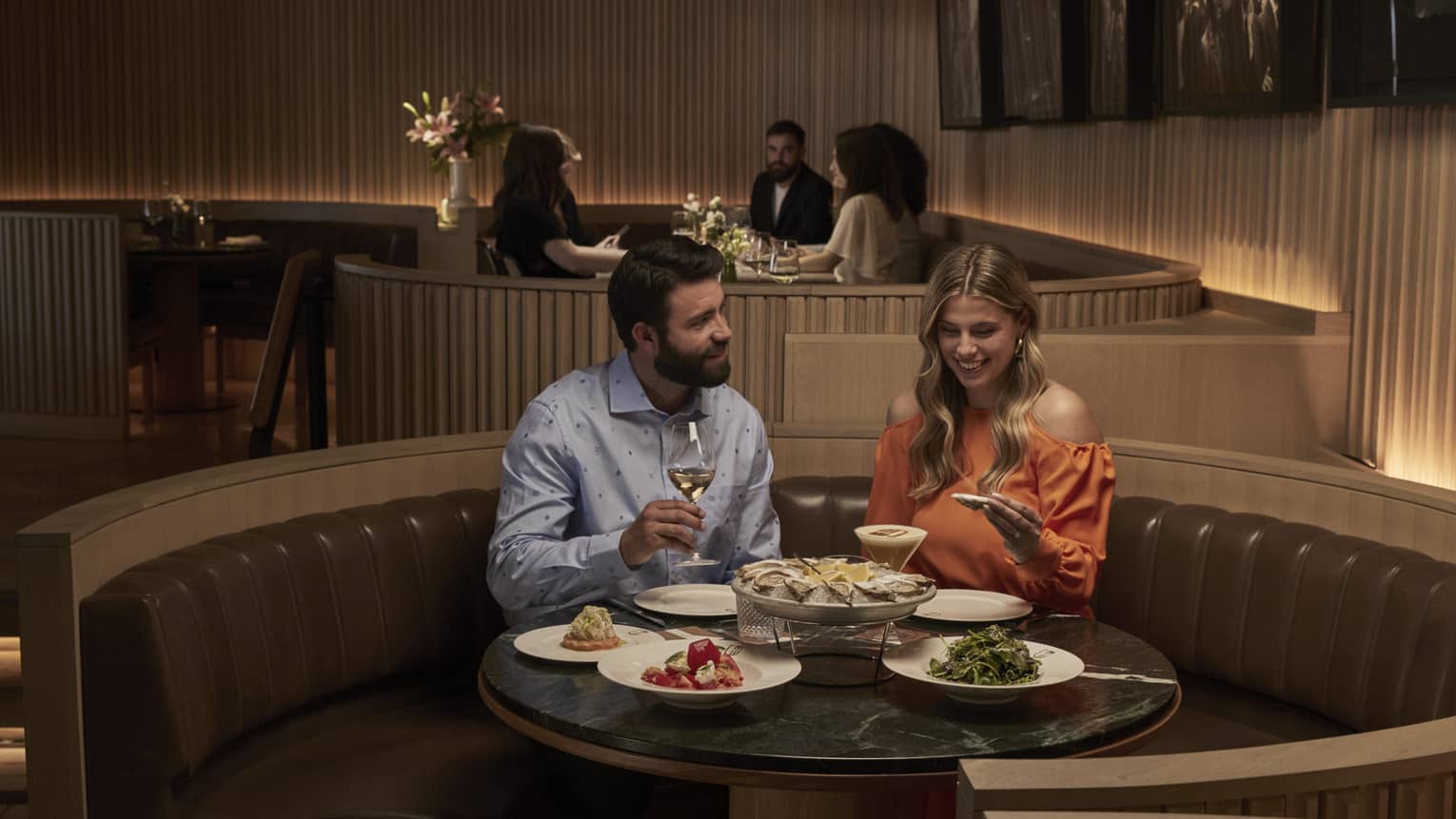 A couple sits at a round table in a leather banquette smiling and eating a plate of oysters and various other side dishes