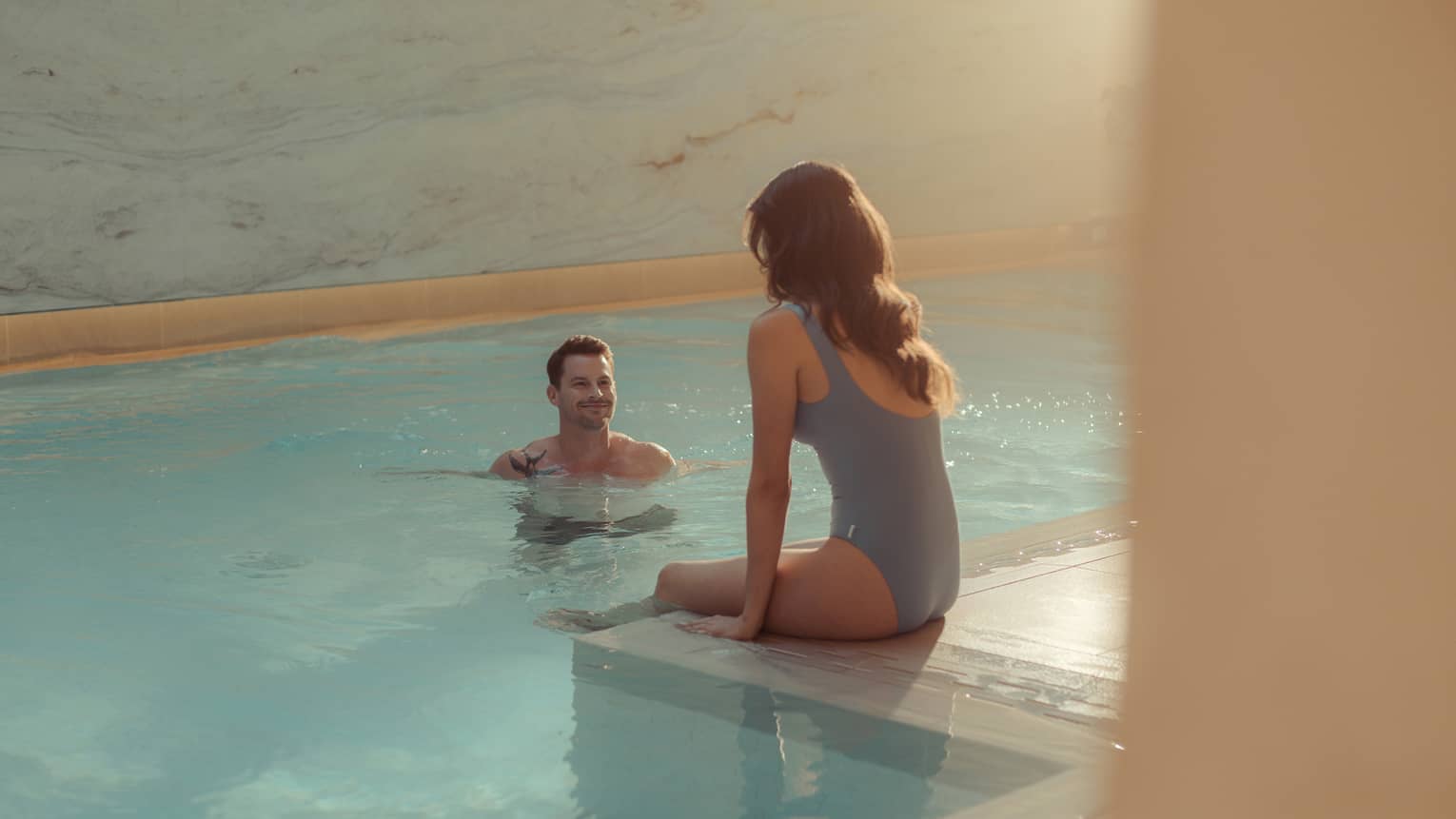 At an indoor pool with marble walls illuminated by natural sunlight, a woman sits on the pool's tiled edge as a man stands in the clear, blue water facing her