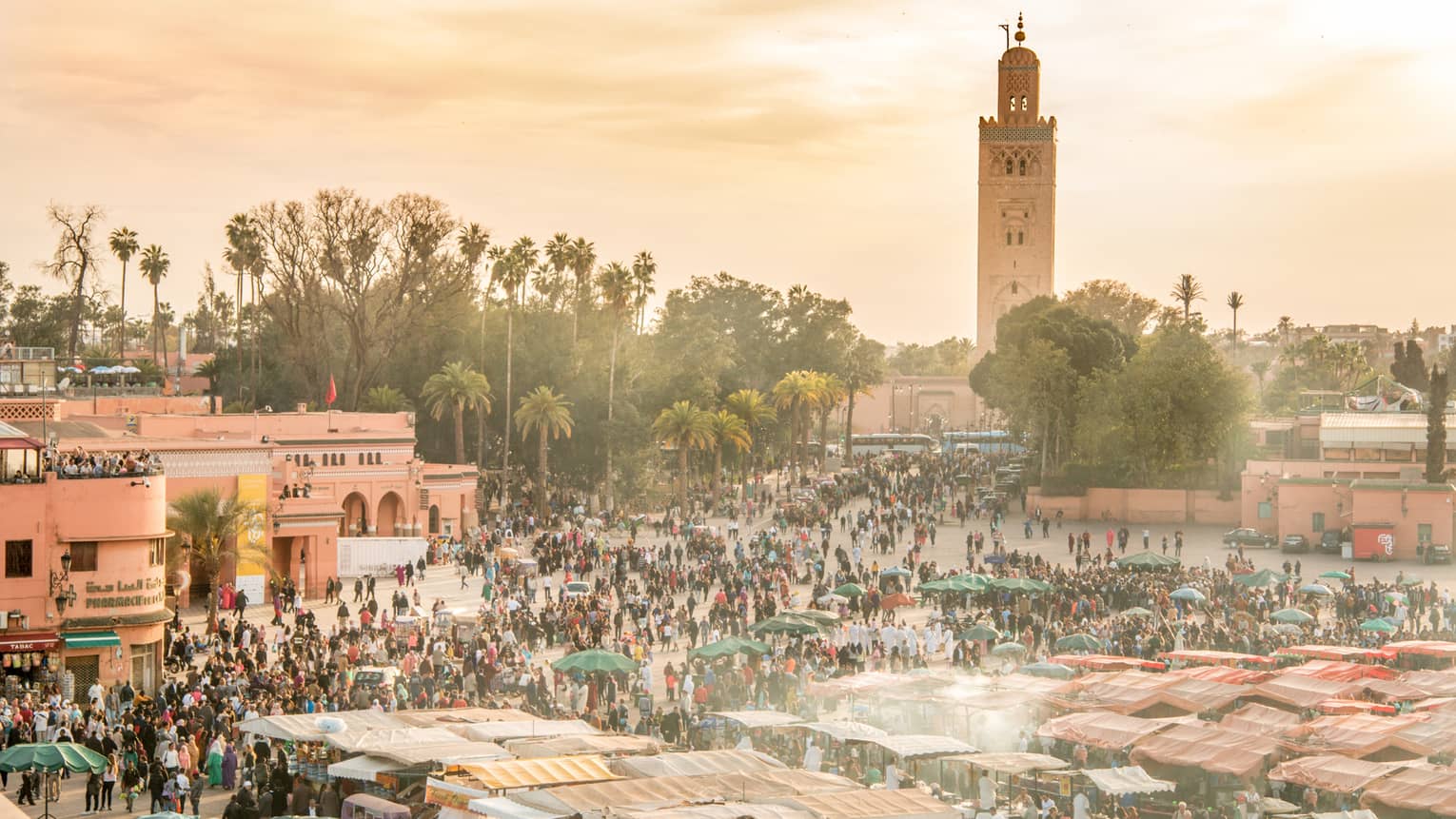 Hazy overhead view of an outdoor market teeming with people, stalls and umbrellas, with trees and a tall minaret at the back.