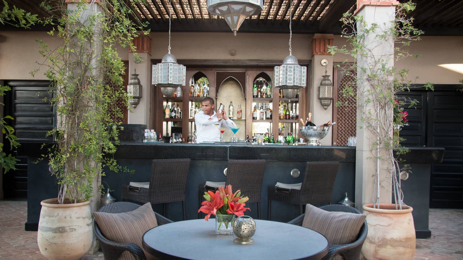 Bartender mixes cocktail in silver shake behind small bar with columns, small table and chair in front