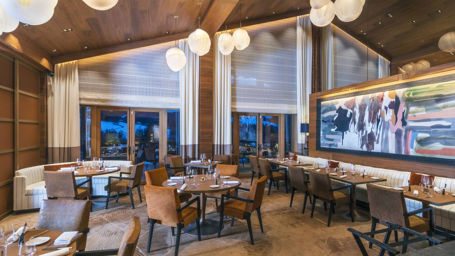 A warm, brightly light, spacious indoor dining area with a peaked ceiling, tables of various sizes and shapes, seating, large floor-to-ceiling windows on one wall, and colourful abstract artwork on another.
