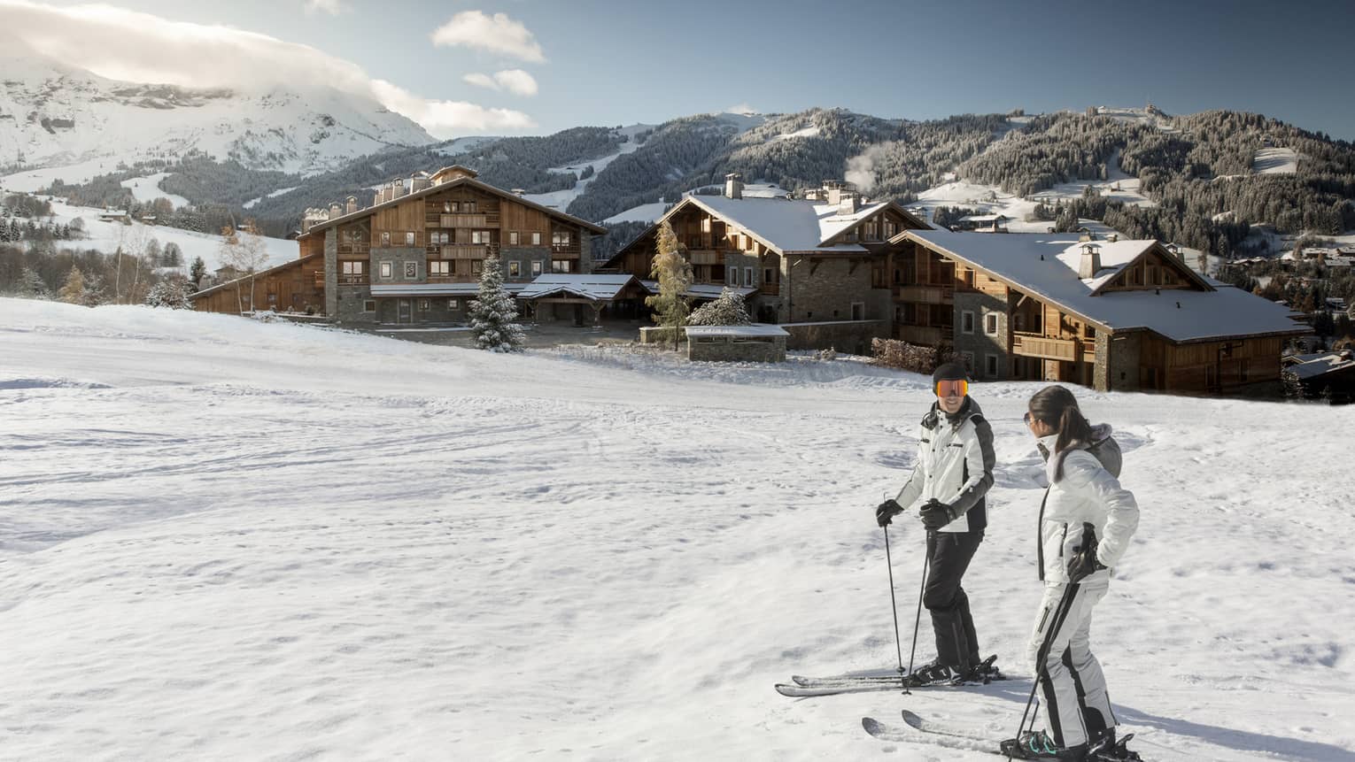 Man, woman on skis at top of snowy hill, Four Seasons Hotel Megeve in background