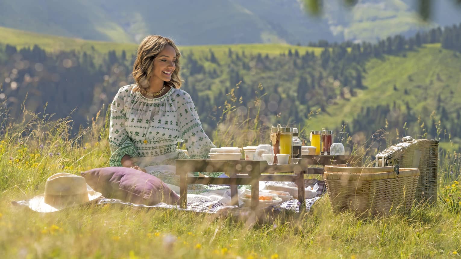 Woman in floral dress sits amid a picnic on a meadow with the Alps in backdrop