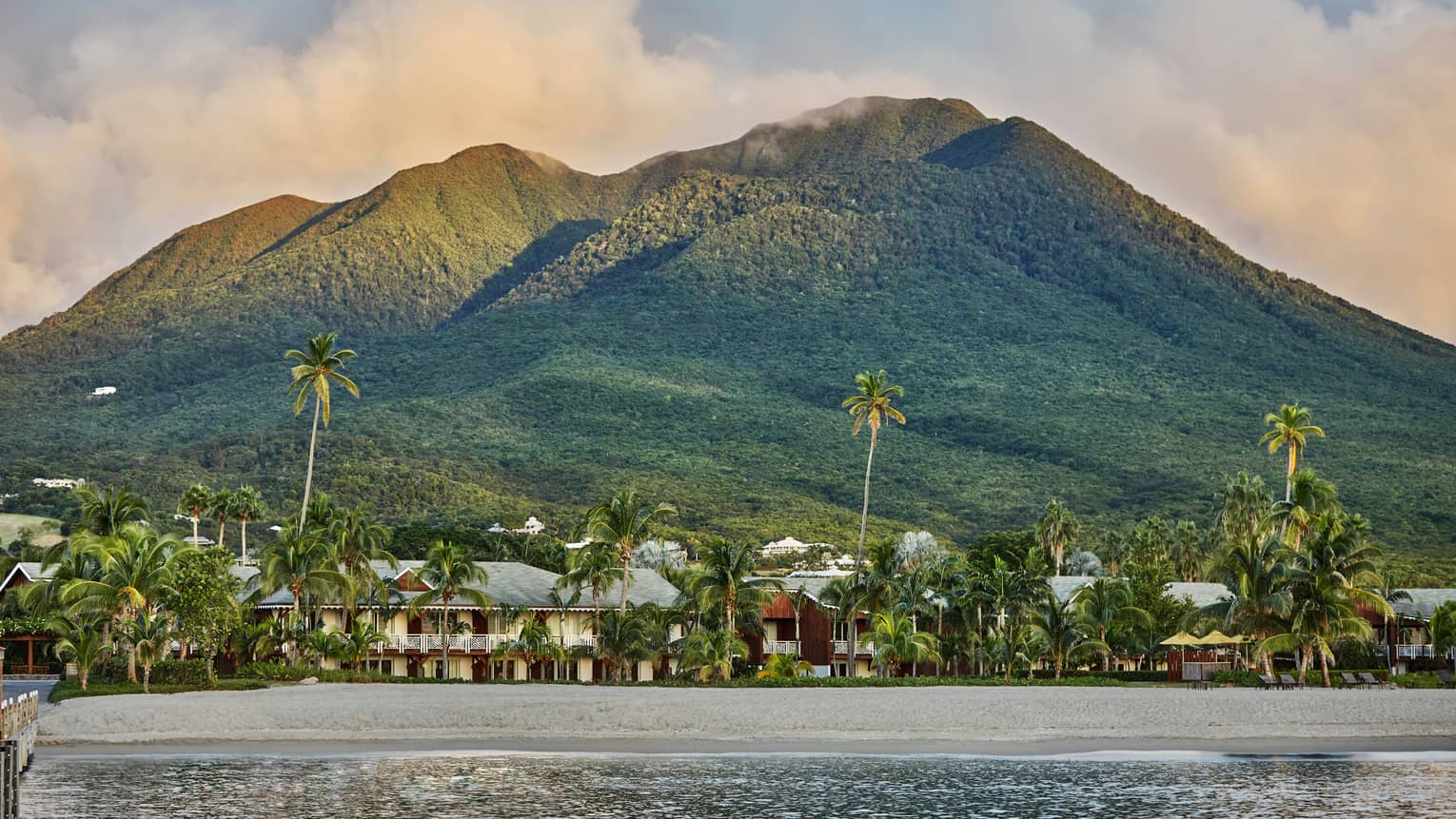 Buildings and palm trees on an ocean shore with a large mountain in the distance.