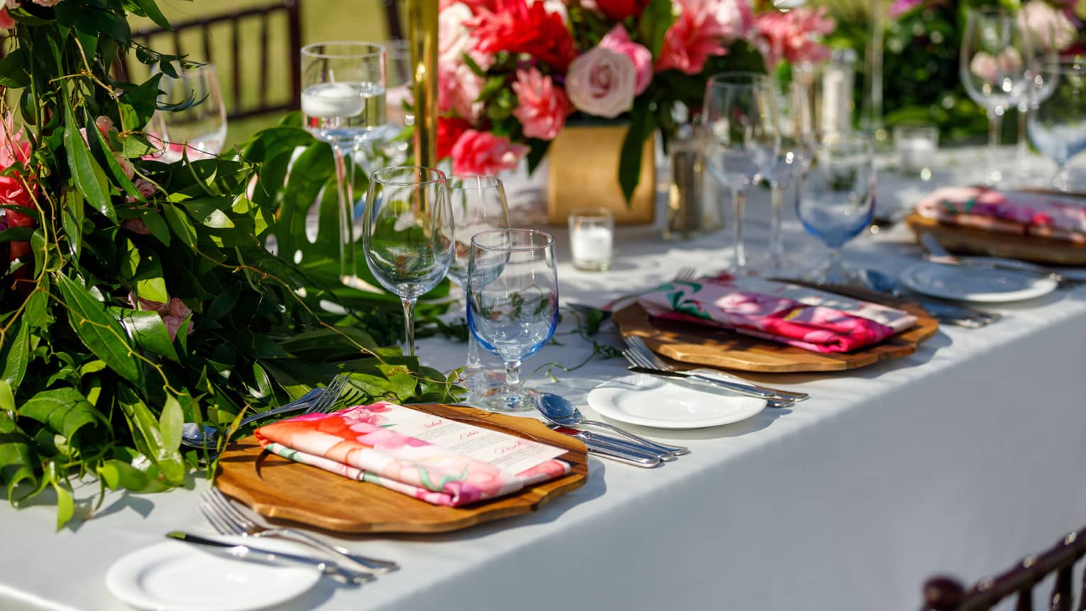 Vibrant table setting with wooden chargers, floral napkins, glassware, pink floral arrangements
