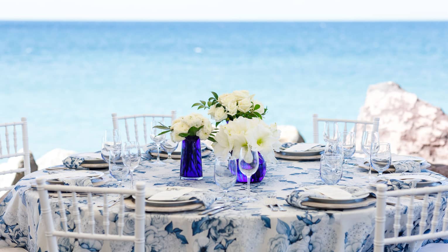 Blue and white round table formally set, five white chairs, white floral centrepieces, on beach