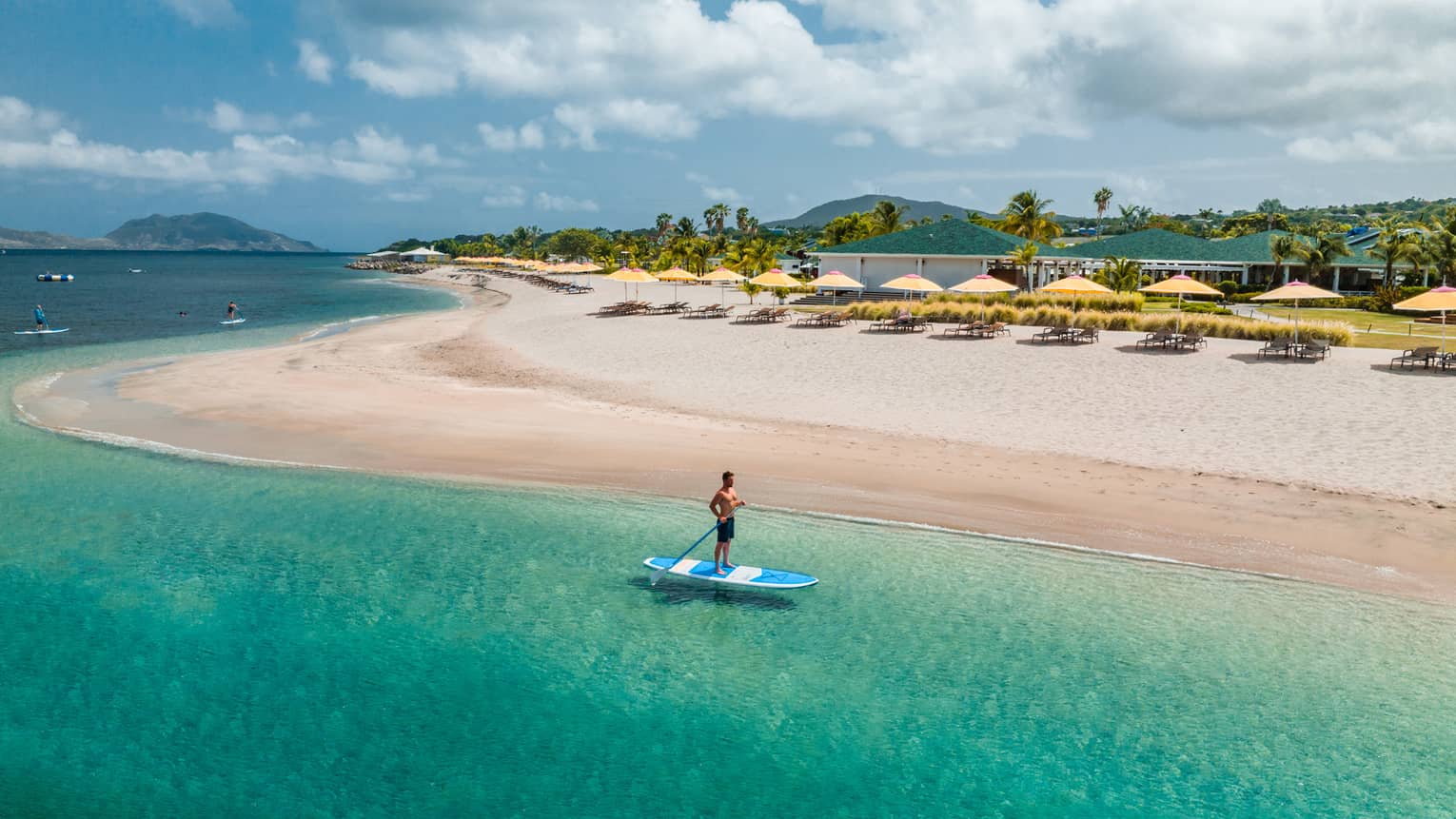 Man stand-up paddleboarding near a shoreline lined with a row of yellow beach umbrellas