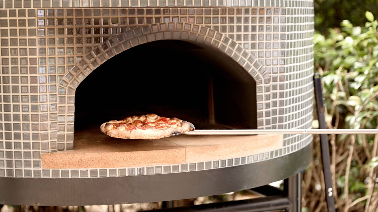Close-up of a wood-fired pizza being pulled from a glass-tiled pizza oven on a long-handled metal pizza peel.