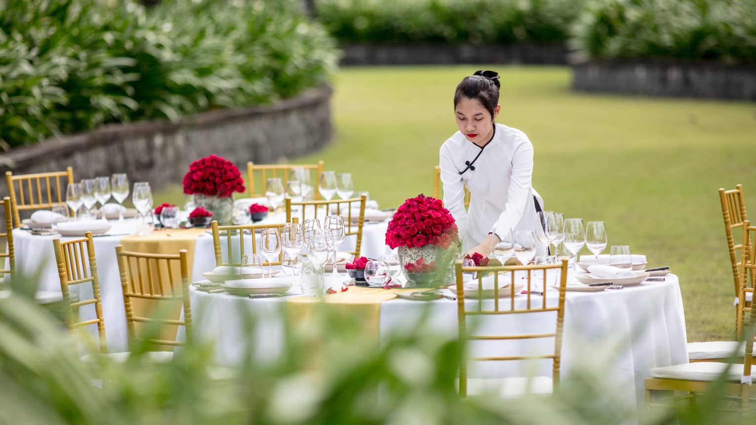 A server sets a beautifully arranged outdoor dining table with gold chairs and red floral centrepieces on a lush lawn.