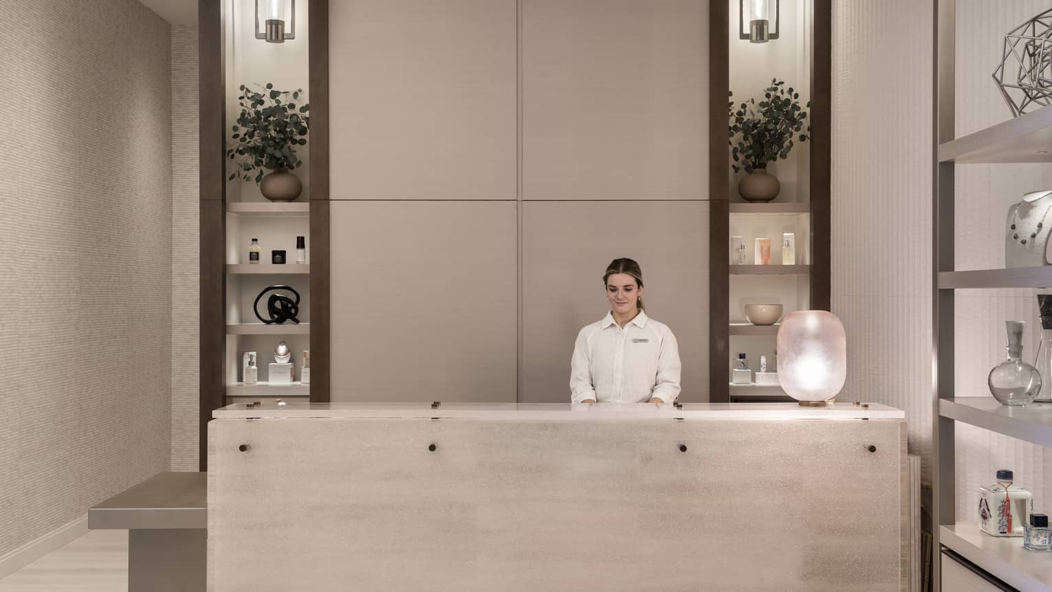 Receptionist in white shirt stands behind white marble spa reception desk with shelving display in backdrop