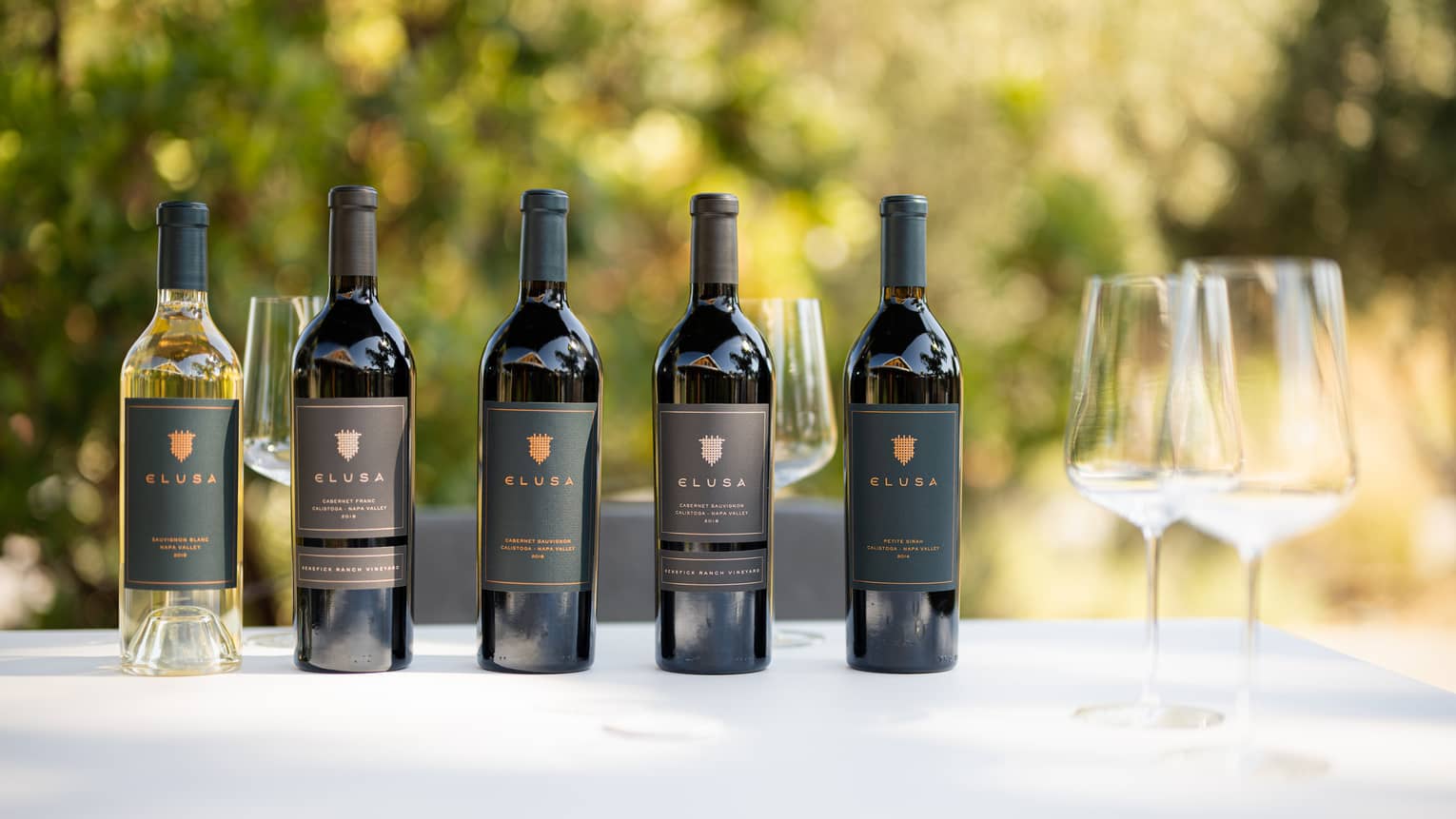A series of wine bottles and two wine glasses sitting on a white table outside.
