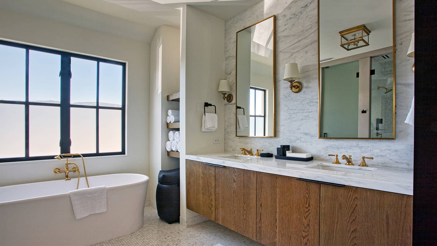 Luxury resort bathroom with a freestanding bathtub, marble double vanity with gold fixtures, mirrors, wall sconces and frosted window for privacy