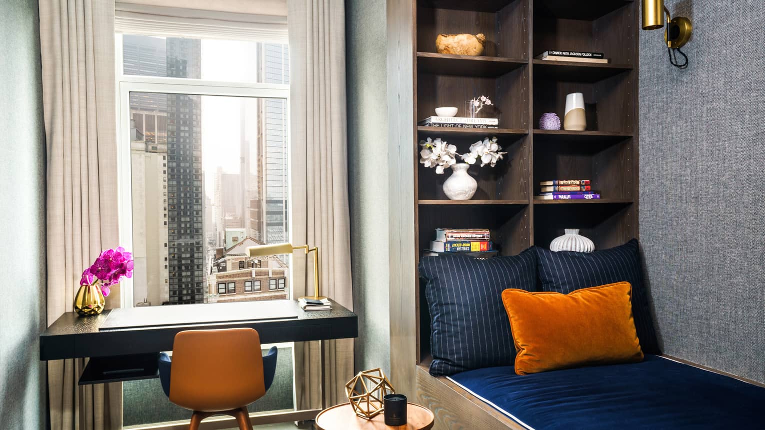 Twin bed with dark blue linens, adjacent to wall with built-in shelves, writing desk, city view