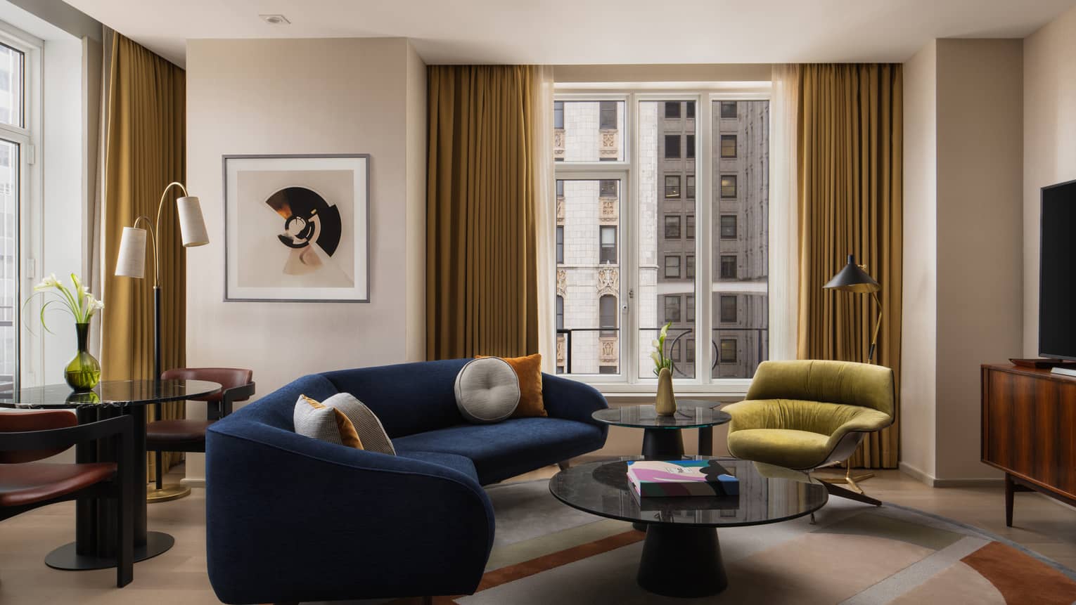 Living room with a navy blue sofa, green lounge chair, round coffee tables, abstract artwork and floor-to-ceiling windows with gold curtains