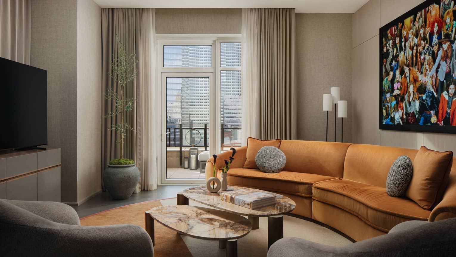 Hotel suite living area with curved orange sofa, coffee tables, gray armchairs, and colourful artwork on the wall, overlooking a balcony with city views