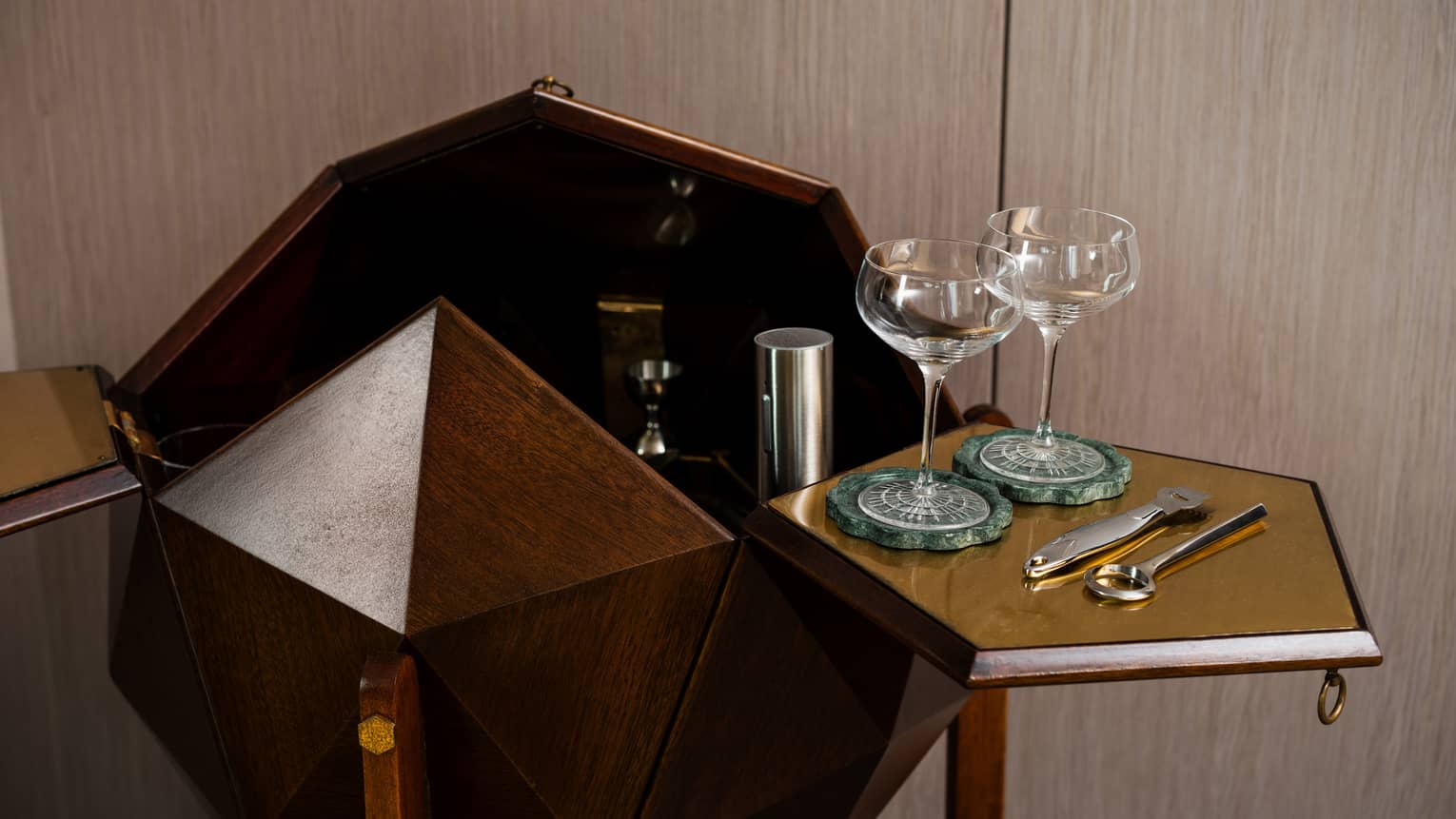 Geometric wooden bar cabinet with open doors, displaying two cocktail glasses on green coasters, a bottle opener, and a cocktail stirrer 
