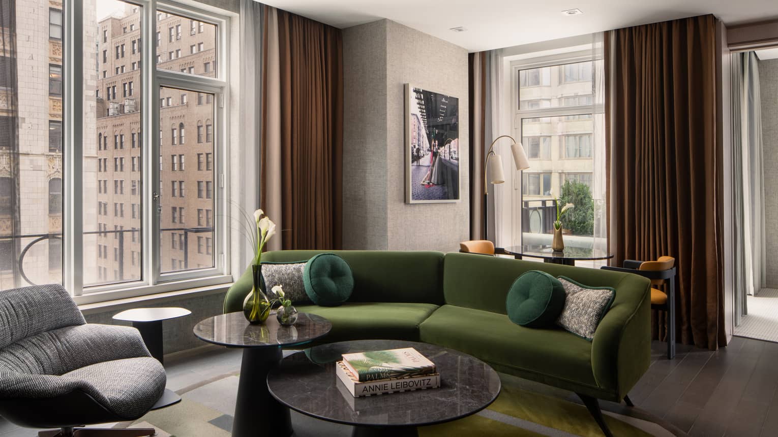 Luxury hotel suite living room with a green velvet sofa, round coffee tables, armchair, wall art, and large windows with views of city buildings