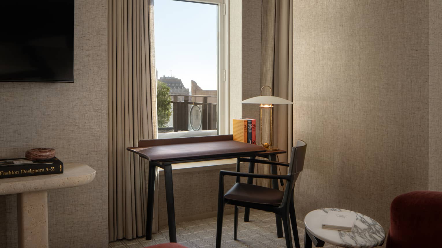 Small desk with a chair and a lamp by a window, adjacent to a round marble table with a book in a cozy hotel room setting