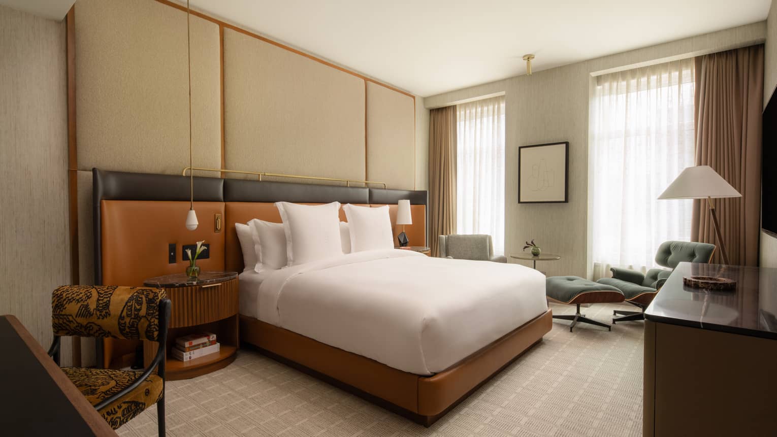 Modern hotel room with king bed, leather headboard spanning the width of the wall, two windows, dresser, desk and chair, and armchair