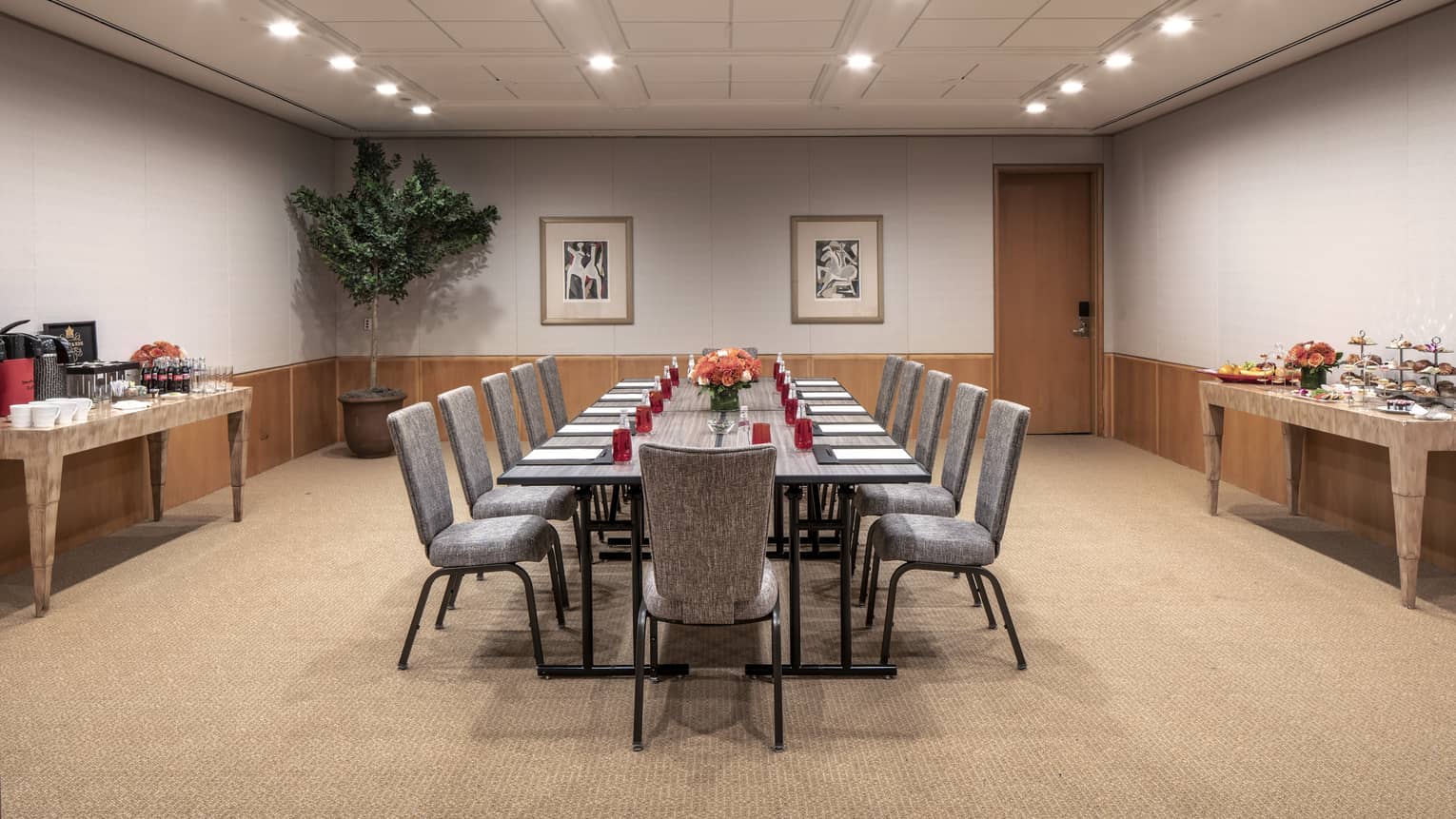 A boardroom with a long rectangular gray table that is set with red water glasses, a black folder, white paper and a orange floral centerpiece.