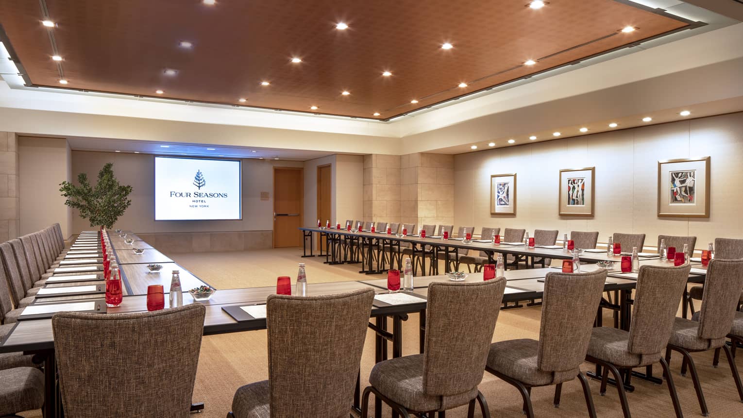 Multiple long, rectangular boardroom tables are placed together to form a "U" shape facing a presentation screen and are set with a black folder, white paper, pen and clear bottles filled with a red soda. 