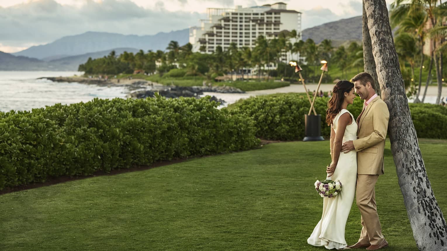 Bride and groom lean against tree on green lawn by ocean, resort 