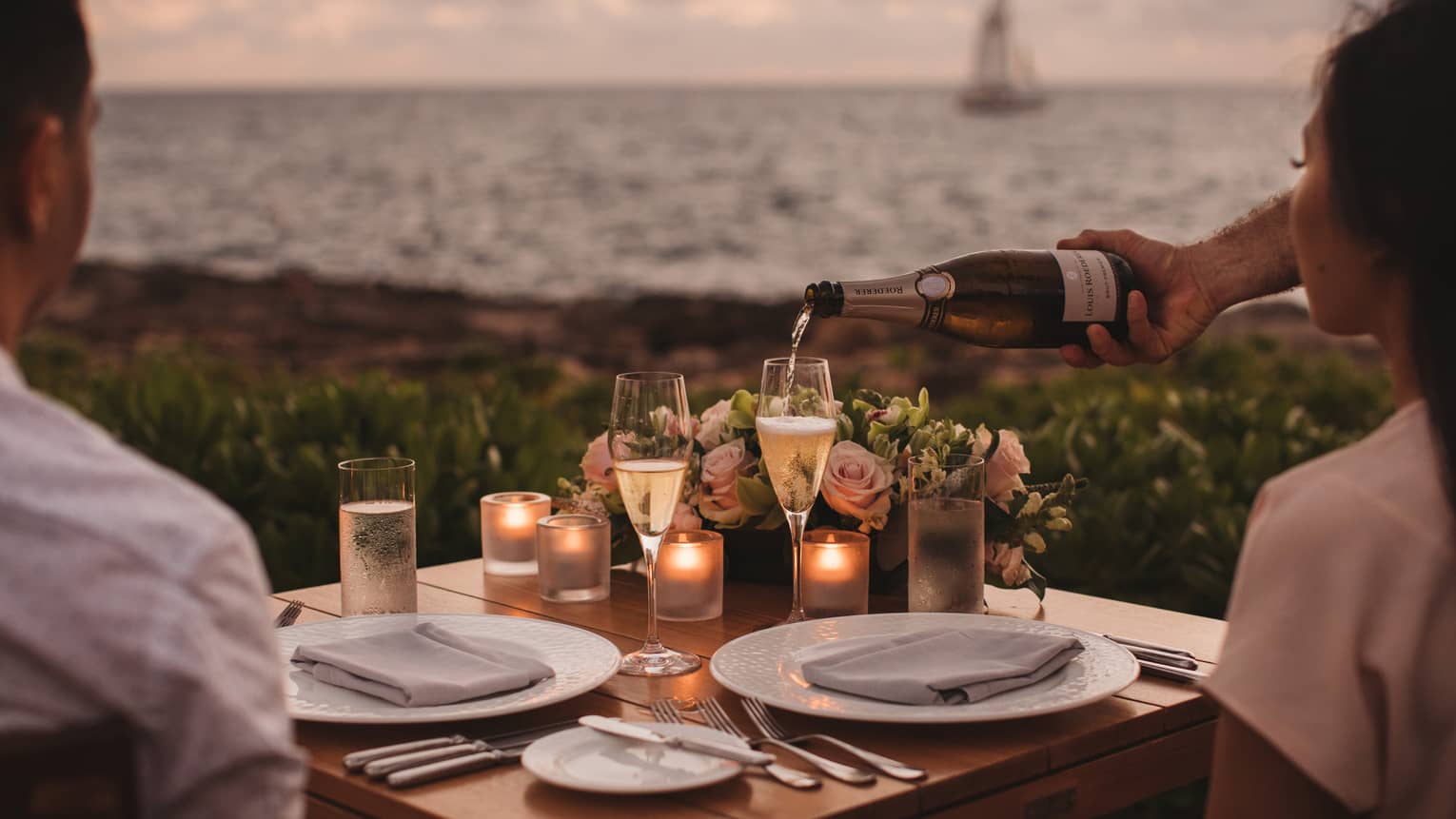 A couple having a private dinner on a coast with a sailboat in the distance.