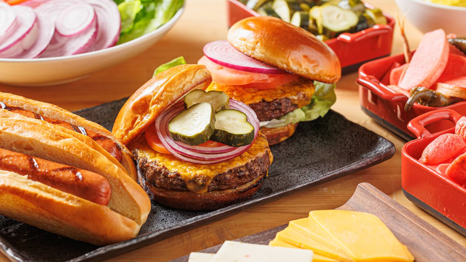 A table holding a variety of food including sliced cheese, a cheeseburger, hotdogs and salad