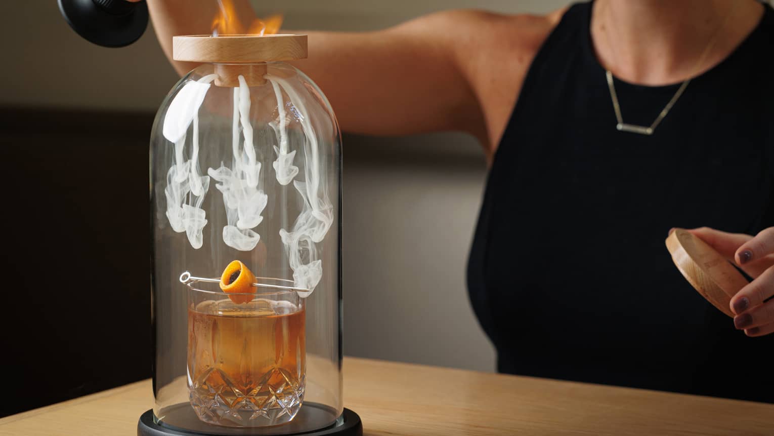 Cocktail set in a glass case with smoke, as a female bartender lights the top with a torch