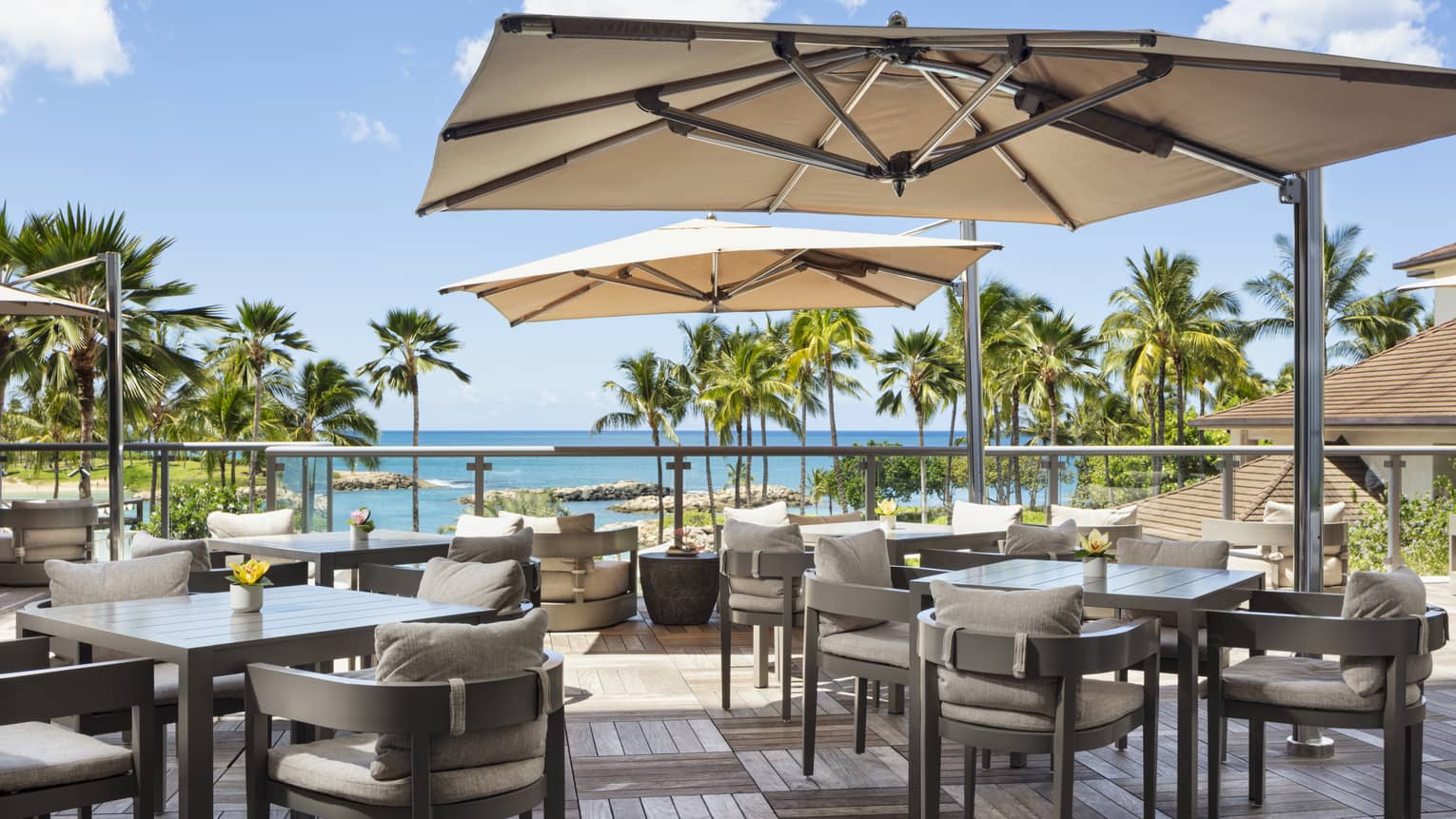 An outdoor dining space has wood deck floors, square tables shaded by large sun umbrellas and chairs with beige cushions and offers a view of palm trees and the ocean.