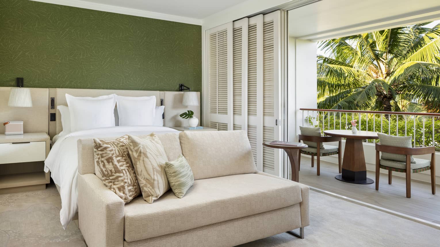 Luxury hotel room featuring a king bed, green accent wall, chaise and private balcony with two chairs and table, looking out to palm trees
