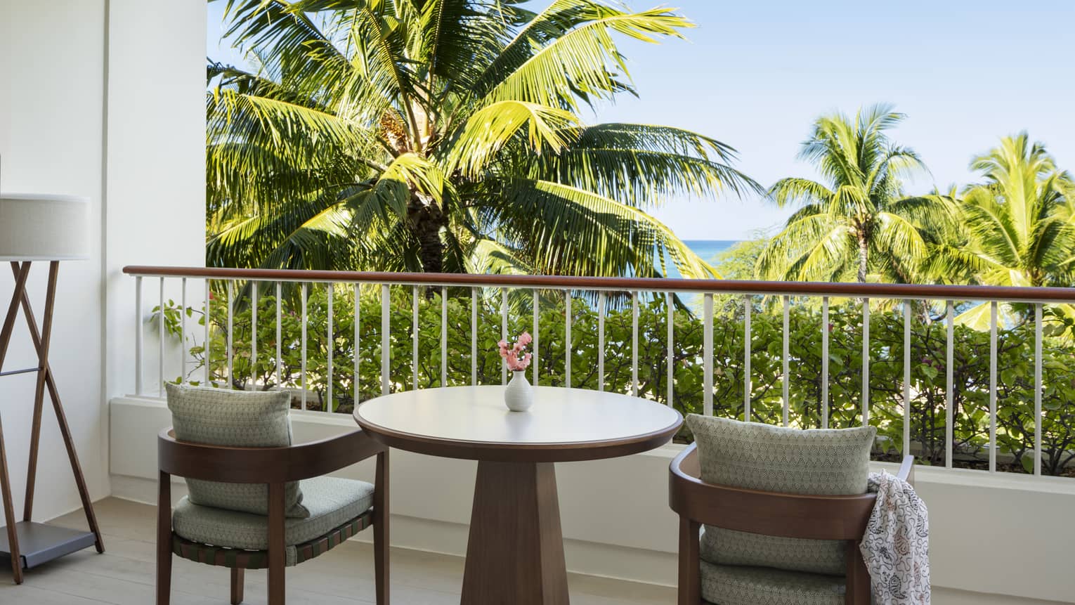 A private hotel balcony with two wooden chairs and a round table, facing palm trees and ocean