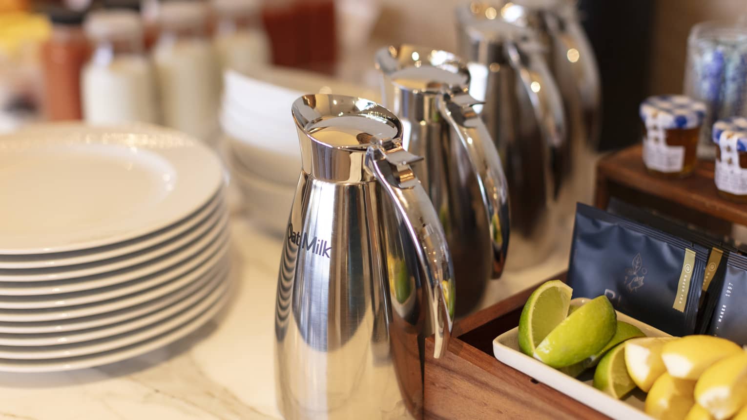 Beverage bar counter containing four silver milk pitchers for coffee, lime and lemon wedges, milk and small jams