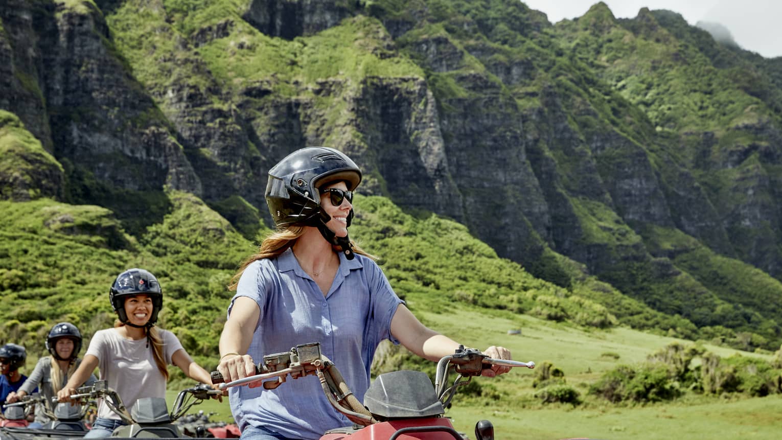 A row of smiling ATV riders wearing black helmets. Behind, a sun-drenched verdant mountain with a mid-layer of craggy rock.