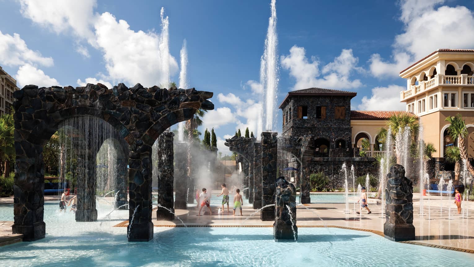 Kids play in water, under sprinklers and stone pillars in the Splash Zone water park