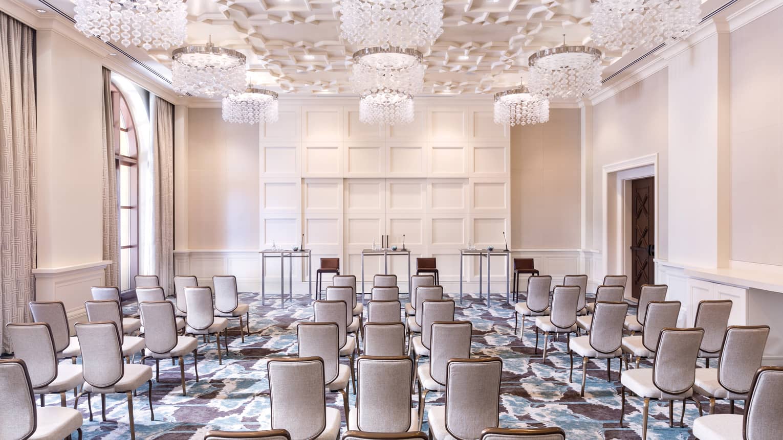A cream colored function room is set up with rows of neutral chairs lined up and facing three tables in the front