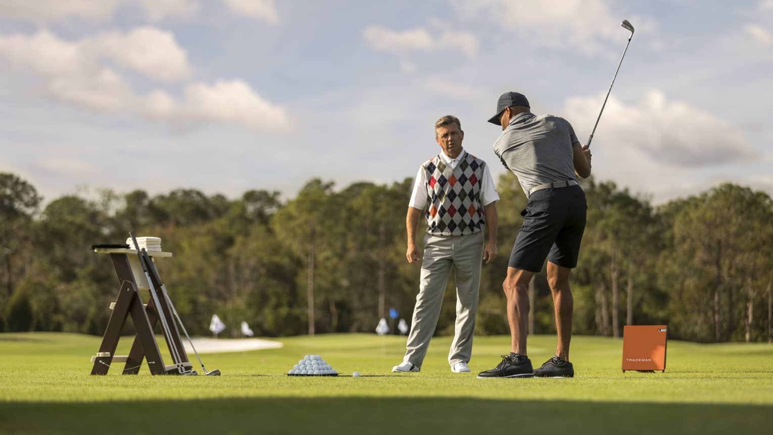 Against clear sky, one golfer looks on as another prepares to swing, a tray of balls and a stand of clubs nearby.